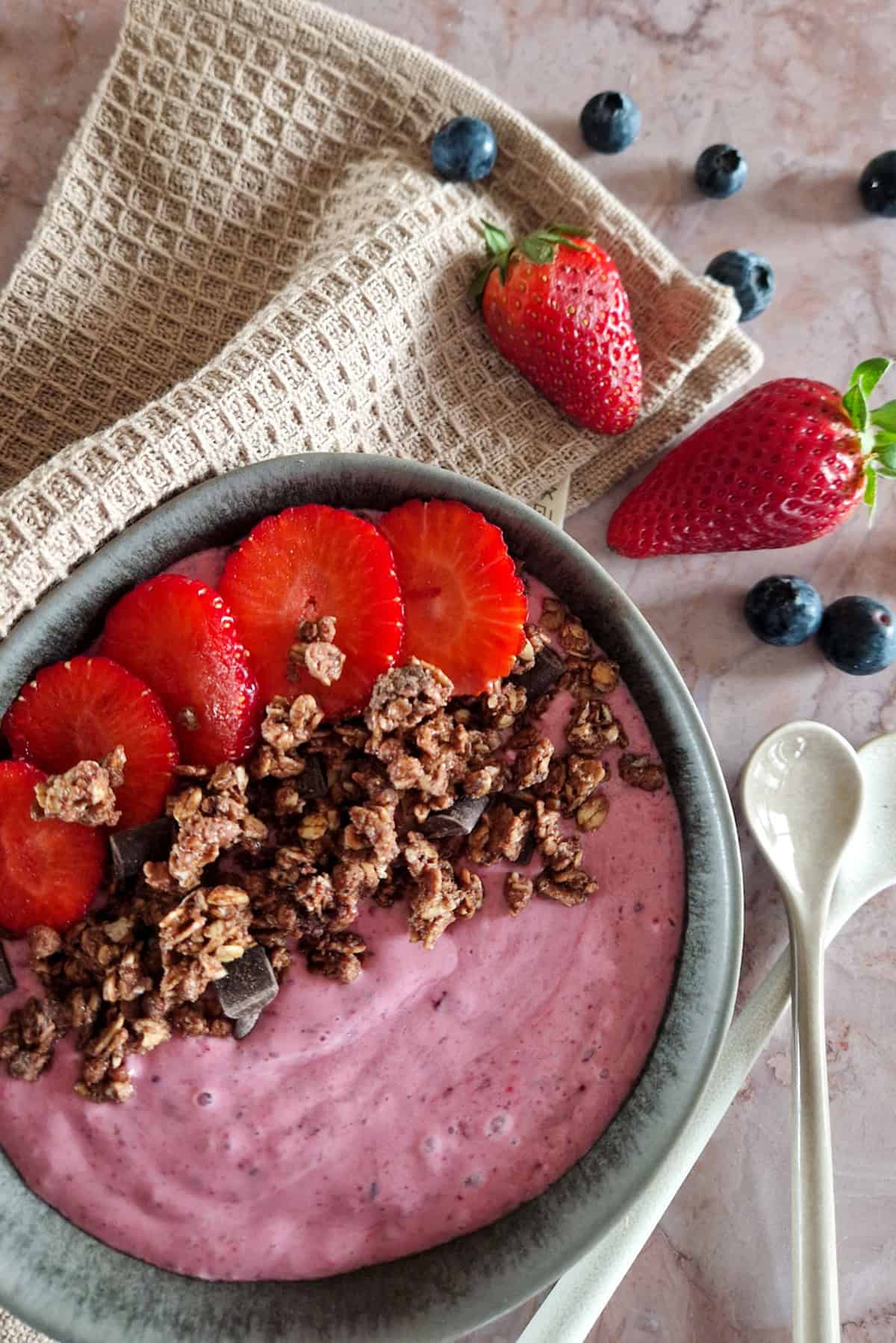 Een close-up van een bosvruchten smoothie bowl van diepvriesfruit, gegarneerd met granola en verse schijfjes aardbei.
