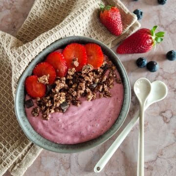 Smoothiebowl met aardbeien en bosvruchten in een donkere kom. De roze smoothiebowl is gegarneerd met granola en verse aardbeien.