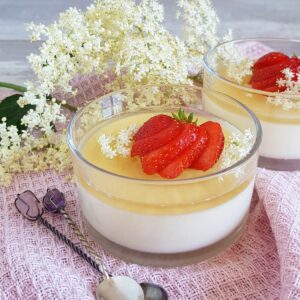 Vlierbloesem panna cotta in een glazen potje. De panna cotta is voorzien van een laagje vlierbloesemjelly en gegarneerd met verse aardbeien.