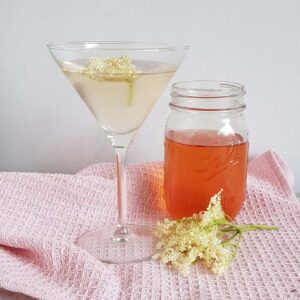 Vlierbloesemlikeur in een cocktailglas. Op de achtergrond een glazen pot met de rest van de likeur, en een vlierbloesemscherm erbij.