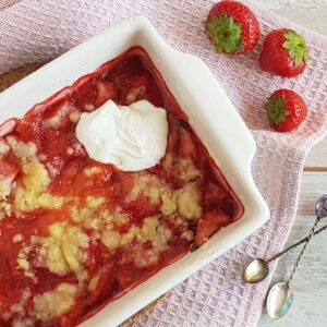 Aardbeiencrumble met een dot vanillemascarpone in een ovenschaal. Er liggen zilveren lepeltjes en verse aardbeien naast de crumble.