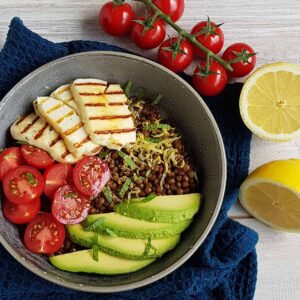 Lunchbowl met linzen en halloumi. De linzensalade is bedekt met schijfjes avocado, gehalveerde tomaatjes en gegrilde halloumi.