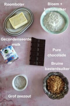 De ingrediënten om zelf caramel chocolade reep te maken: bloem, boter, suiker, gecondenseerde melk, pure chocolade en grof zeezout. De ingrediënten zijn uitgestald op een ondergrond met marmerpatroon.