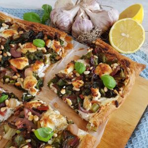 Plaattaart met snijbiet en geitenkaas. De plaattaart ligt op een houten serveerplank en is in stukken gesneden.