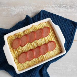 Spitskool stamppot ovenschotel met plakjes rookworst gegarneerd. De schotel staat op een donkerblauwe doek.