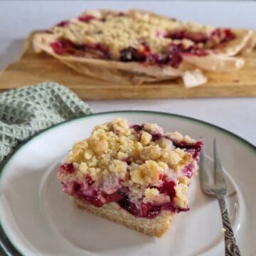 Pruimen plaatkoek op een gebaksbordje met een zilveren vorkje. Op de achtergrond een bakblik met de rest van de pruimentaart met kruimeldeeg.