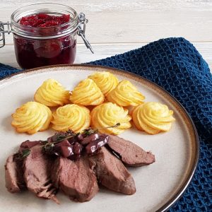 Hertenbiefstuk, in plakjes gesneden en gegarneerd met champignonsaus en verse tijm. Er liggen pommes duchesses bij.