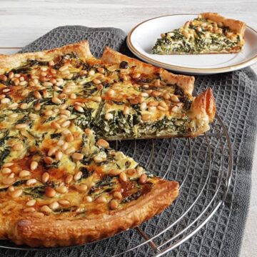 Spinaziequiche met oude kaas op een ovenrek. De quiche is rijkelijk bestrooid met pijnboompitten en er is een punt uitgesneden zodat je de vulling van spinazie en oude kaas goed kunt zien.