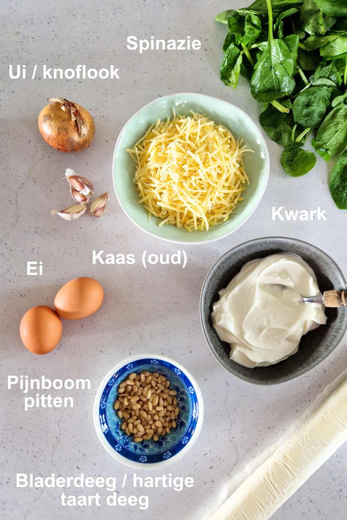 De ingrediënten voor een spinaziequiche: deeg, spinazie, kaas, kwark, eieren, knoflook, ui en pijnboompitten.