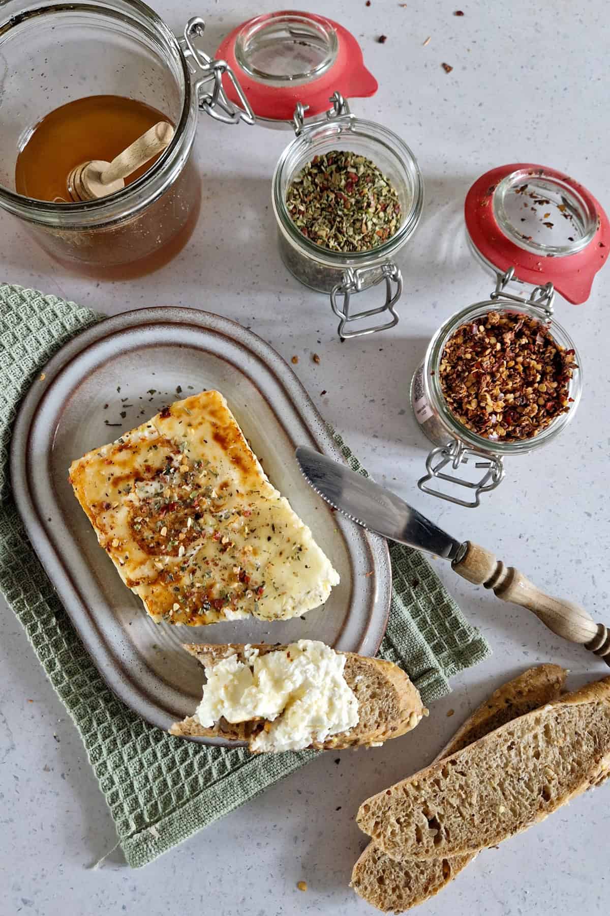 Een Grieks recept voor gebakken feta (saganaki). De feta is op een broodje gesmeerd en er omheen staan de smaakmakers van het recept: honing, oregano en chilivlokken.