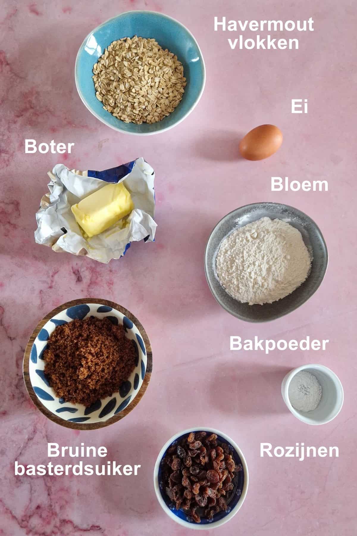 De ingrediënten voor havermoutkoekjes met rozijnen, uitgestald op een roze-marmeren ondergrond.