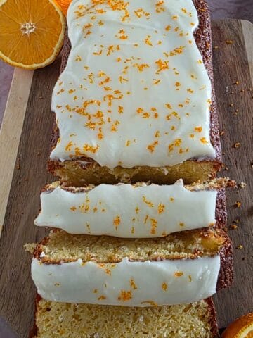Een in plakjes gesneden sinaasappelcake op een houten serveerplank. De cake is gegarneerd met een witte frosting en sinaasappelrasp.