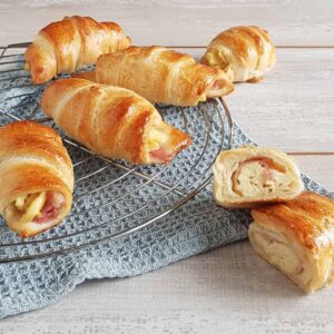 Croissantjes met ei en ontbijtspek op een ovenrek. Eén van de croissants is opengesneden zodat je de vulling van roerei en spek goed kunt zien.