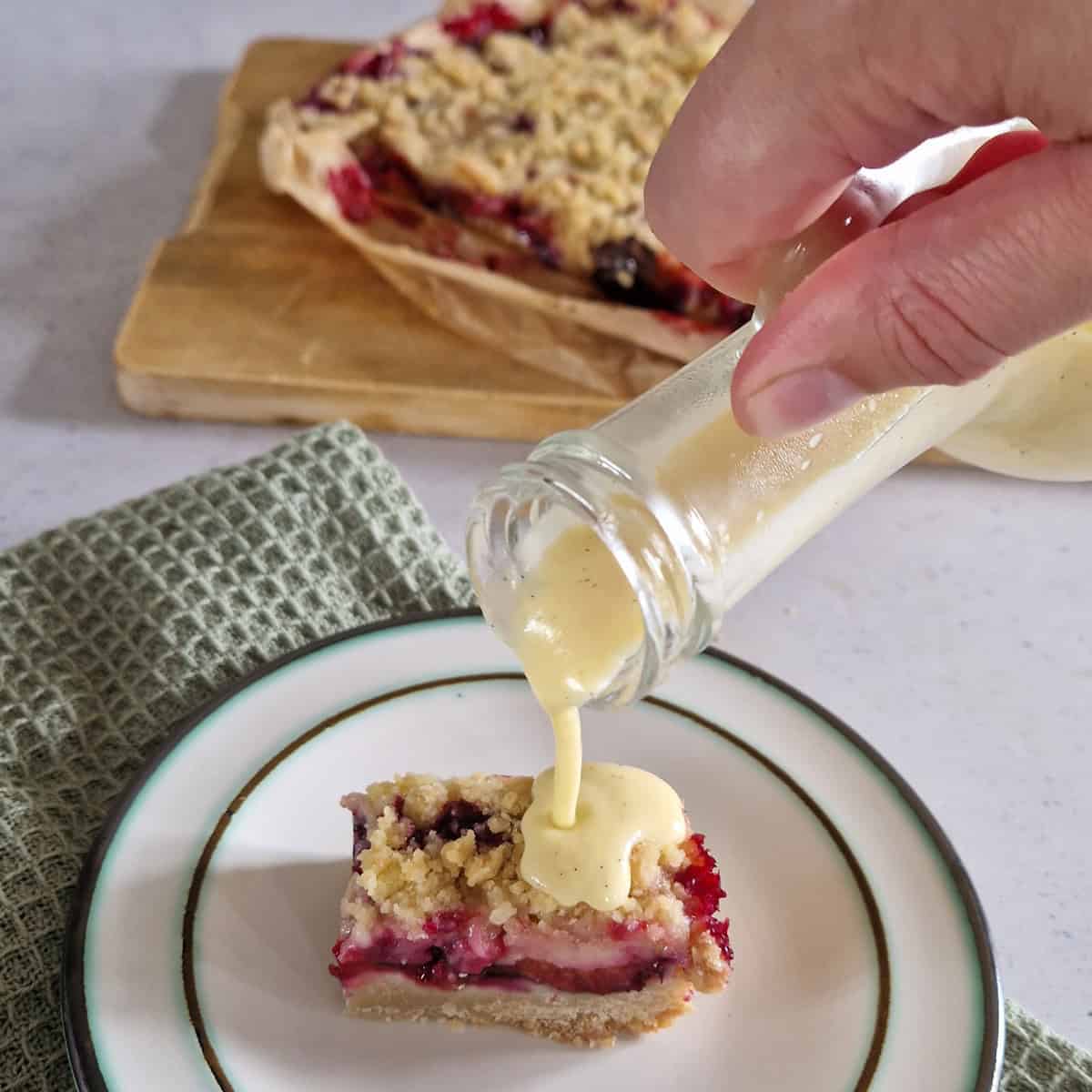 Vanillesaus in een schenkkannetje, een hand schenkt de saus over een stukje taart.