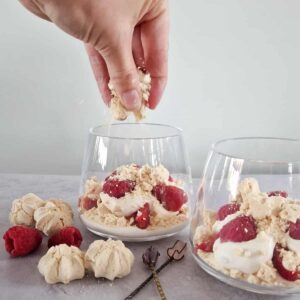 Een hand verkruimelt meringue boven een Eton Mess met frambozen.