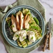 Gegrilde courgette met kip, pesto en een bol mozzarella. Het gerecht ligt op een ouderwets bordje, dat op een lichtgroene stoffen doek staat.