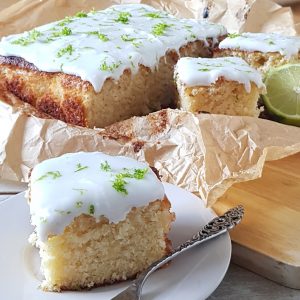 Kokoscake met limoen, uit de cake is een stuk gesneden dat op een bordje ligt. De kokoscake heeft een dikke laag witte frosting met limoen.