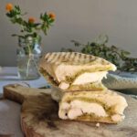 Close-up van een panini met kip en pesto. De panini is gehalveerd en ligt op een houten plank, er staat een glazen vaasje met oranje bloemen op de achtergrond ter decoratie.