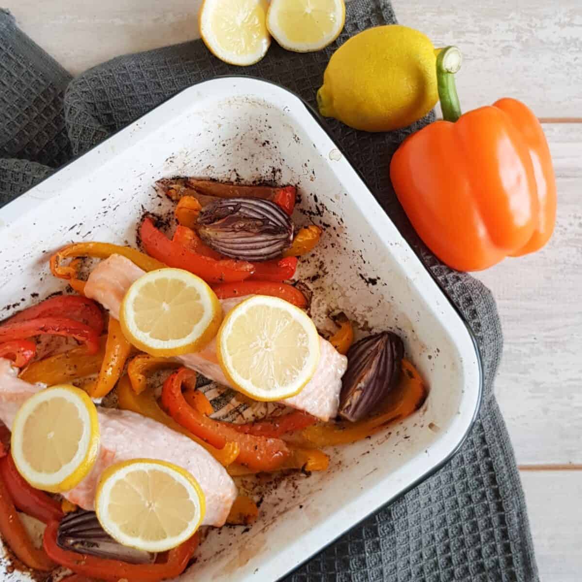 Traybake met zalm en paprika, in de ovenschotel zijn verschillende soorten paprika en rode ui gebruikt. Er liggen twee moten zalm op, waar schijfjes citroen op liggen.