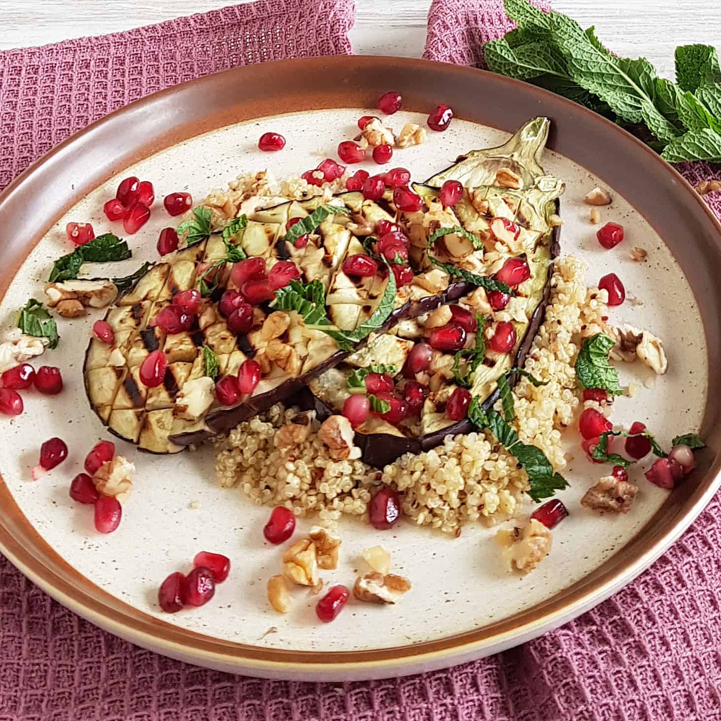 Gegrilde aubergine met quinoa, granaatappel, gehakte noten en verse munt. De aubergine is in zijn geheel in dikke plakken gesneden en kruislings ingesneden.