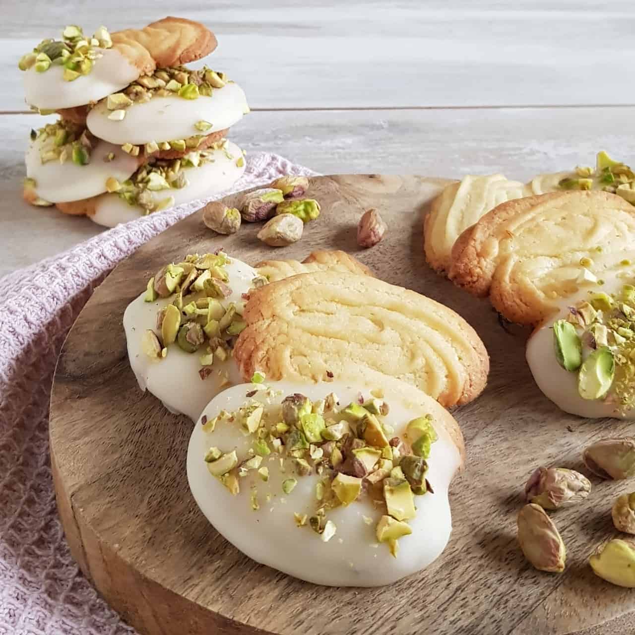 Spritsen met witte chocolade en fijngehakte pistache. De koekjes liggen op een houten serveerplank en zijn deels op elkaar gestapeld.