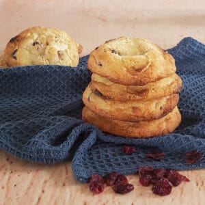 Cranberry koekjes met witte chocolade, op elkaar gestapeld. De koekjes staan op een blauwe doek en er liggen gedroogde cranberries voor.