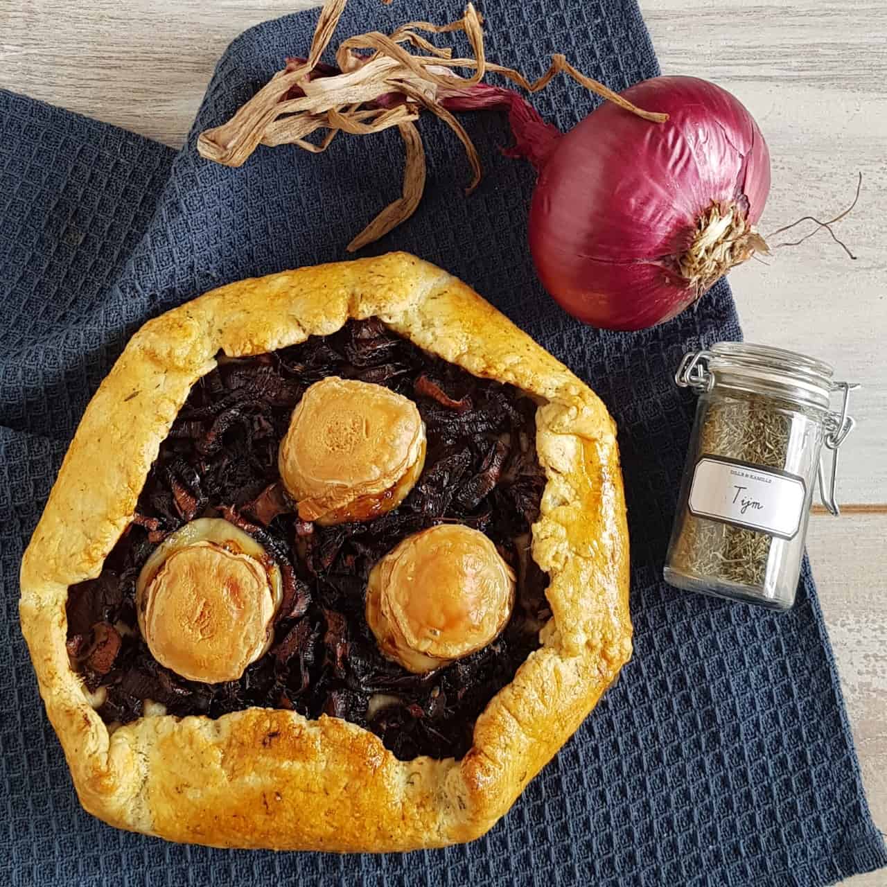 Hartige galette met rode ui en geitenkaas. Er ligt een glazen potje met gedroogde tijm naast de geitenkaas galette.