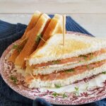 Club Sandwich zalm op een bord met een ouderwetse, decoratieve rand. De Club sandwich is belegd met kruidenkaas en zalm. De helft van de sandwich ligt op een kant zodat je de laagjes goed kunt zien.