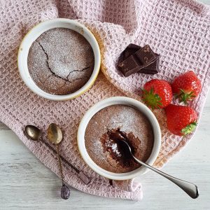 Chocolade lava cake in twee potjes. In één van de potjes is een lepel gestoken, zodat je de vloeibare binnenkant kunt zien. Naast de bakjes lavacake liggen aardbeien en chocolade.