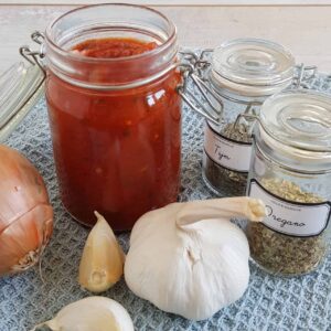 Pizzasaus in een potje. Er omheen staan ingrediënten om zelf pizzasaus te maken: knoflook, ui, oregano, tijm.