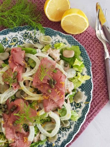 Vitello tonnato maaltijdsalade op een groot bord met blauwe rand. De salade is rijkelijk gevuld met rosbief, venkel, bonen en kappertjes, en gegarneerd met venkelloof.