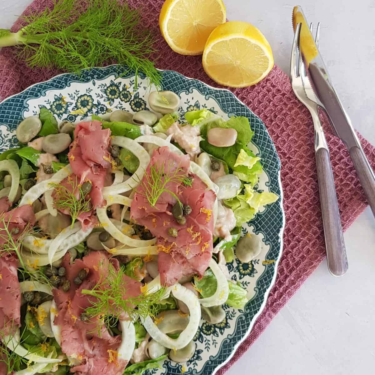 Vitello tonnato maaltijdsalade op een groot bord met blauwe rand. De salade is rijkelijk gevuld met rosbief, venkel, bonen en kappertjes, en gegarneerd met venkelloof.