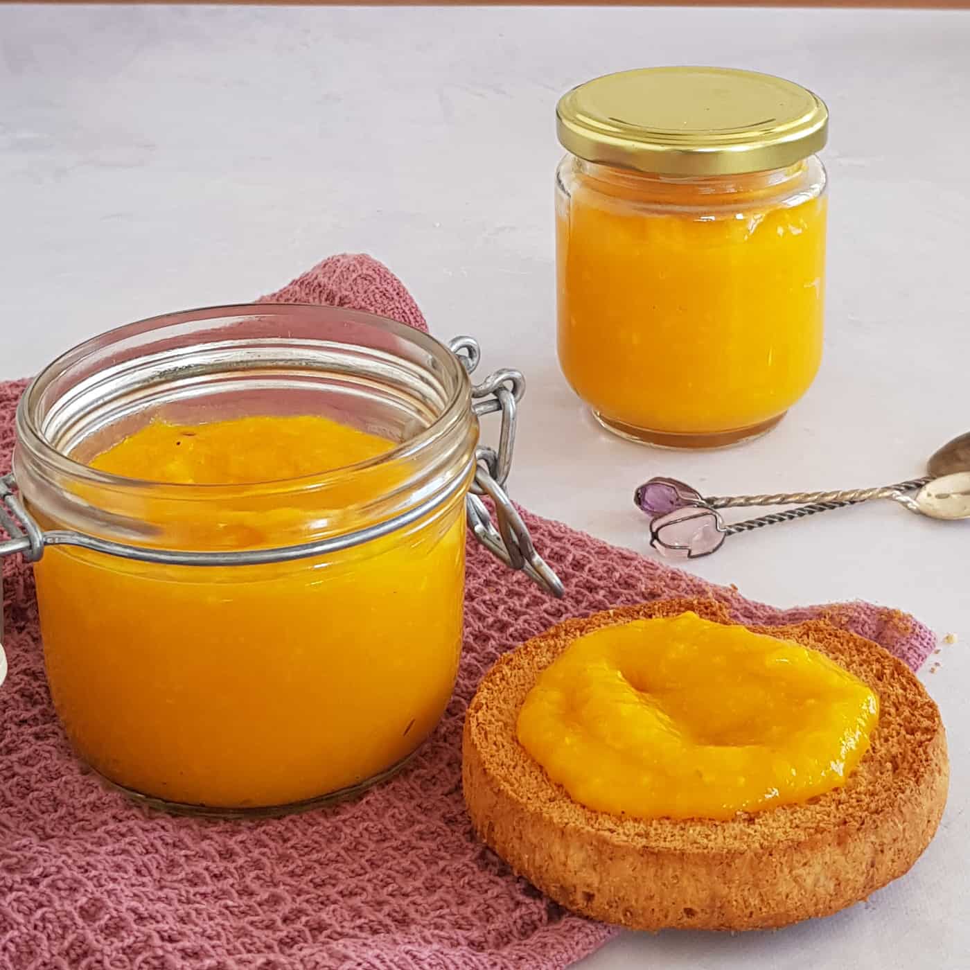 Mango pompoenpuree op een beschuitje. Naast de beschuit en erachter staan twee weckpotjes die gevuld zijn met mango-pompoenpuree.