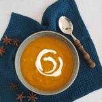 Pompoen-wortelsoep in een kom, met een swirl van yoghurt erin. De soepkom staat op een donkerblauwe doek waar enkele steranijs op liggen.