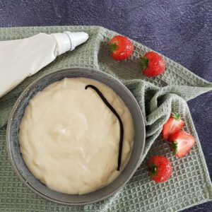 Banketbakkersroom maken met echte vanille. De room zit in een grijze kom en is gegarneerd met een vanillestokje. Er ligt een spuitzak naast en wat verse aardbeien.