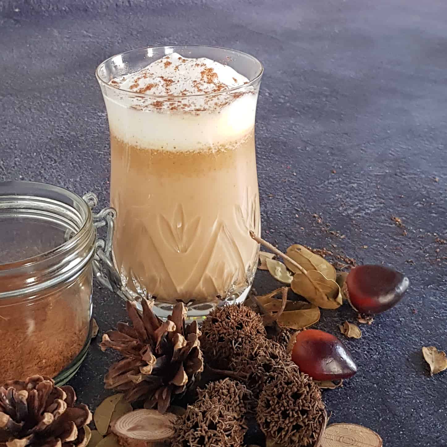 Pumpkin spice latte in een hoog glas. Voor het glas liggen herfstdecoraties zoals kastanjes en dennenappels.