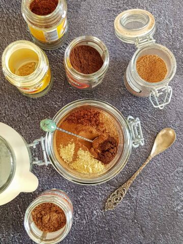 Pumpkin spice mix in een glazen pot met een lepeltje erin. De verschillende specerijen zijn nog niet door elkaar geroerd. Om de pot heen staan kleinere potjes met verschillende soorten ingrediënten.