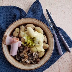 Raclette aardappels met ham en augurk en champignons op een ouderwets bruin bord. Er ligt bestek naast en het bord staat op een donkerblauwe doek.