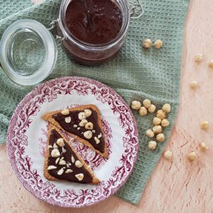 Zelf hazelnootpasta maken met als resultaat deze pot met hazelnootpasta en twee crackers die besmeerd zijn met de pasta. De crackers zijn bestrooid met grof gehakte hazelnoten.