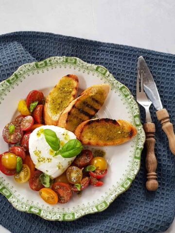 Mozzarellasalade met knoflookbrood en verschillende kleuren snoeptomaten. Het gerecht is gegarneerd met een pestodressing en verse basilicum.