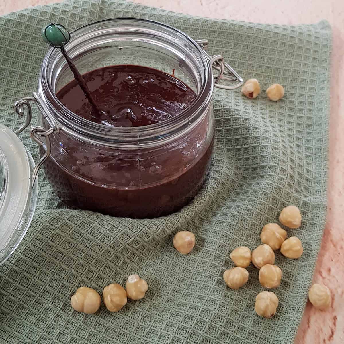 Een pot zelfgemaakte Nutella met een lepeltje erin, er omheen wat hazelnoten ter decoratie.