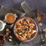 Pompoenpoffertjes met stukjes banaan, blauwe bessen en pittenmix. Er staat een pot notenpasta naast de poffertjes met pompoen.