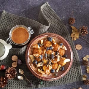 Pompoenpoffertjes met stukjes banaan, blauwe bessen en pittenmix. Er staat een pot notenpasta naast de poffertjes met pompoen.