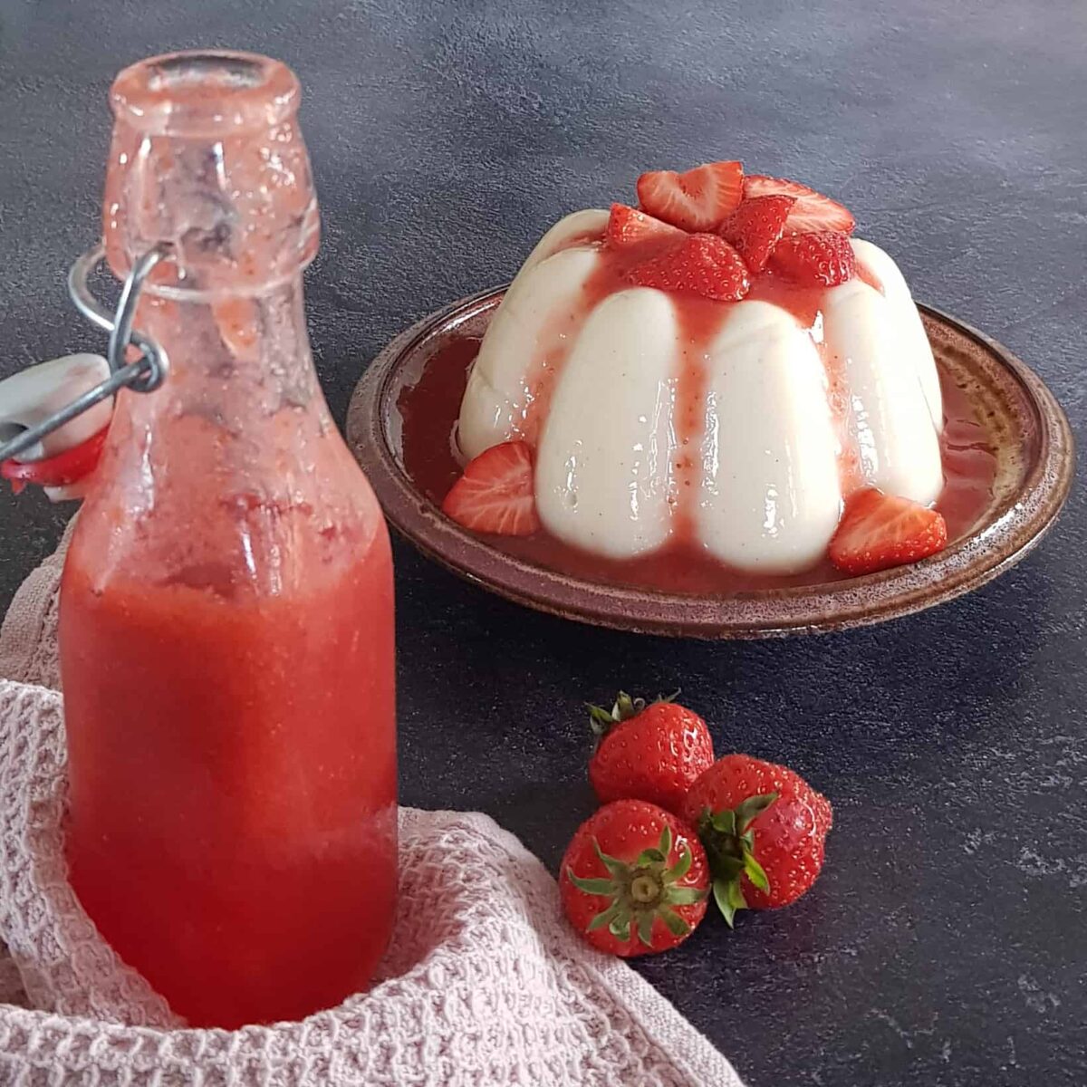 Zelf aardbeiensaus maken met als resultaat een weckfles vol verse aardbeien saus. Op de achtergrond staat een pudding met aardbeien.