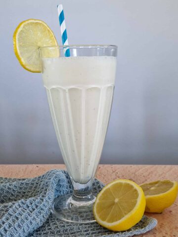 Citroen milkshake in een hoog glas. Het glas staat op een doek en er ligt een gehalveerde citroen bij.