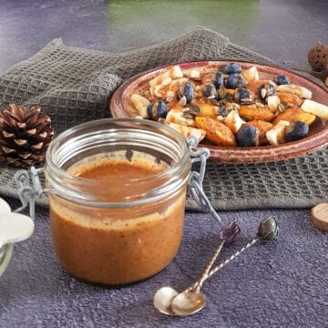 Amandelpasta in een glazen weckpot met zilveren lepeltjes ervoor. Op de achtergrond een bord poffertjes waar de amandelpasta overheen is gesprenkeld.