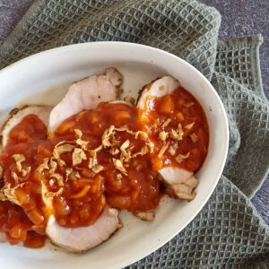 Babi pangang recept: het eindresultaat ligt in een witte ovenschaal die op een doek staat. Over het varkensvlees is een zoete tomatensaus geschept die bestrooid is met gebakken uitjes.