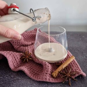 Een hand schenkt zelfgemaakte kaneellikeur uit een fles in een glas. Het glas staat op een paarse doek met steranijs en een kaneelstokje ernaast.