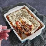 Pompoenlasagne recept: een hand houdt een lepel omhoog met een grote schep van de lasagne, zodat de verschillende laagjes zichtbaar zijn.