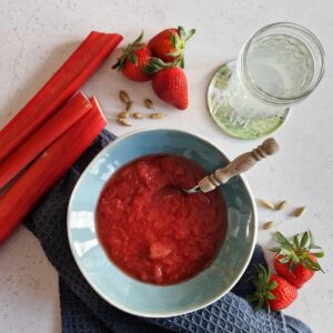 Een kom rabarbercompote met aardbeien. Er staat een lepel met een houten handvat in de kom.